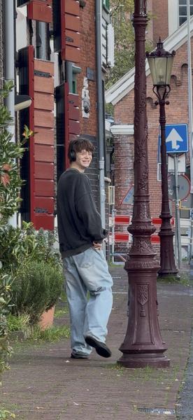 A young person wearing headphones and casual clothes walks along a brick sidewalk, turning to smile at the camera